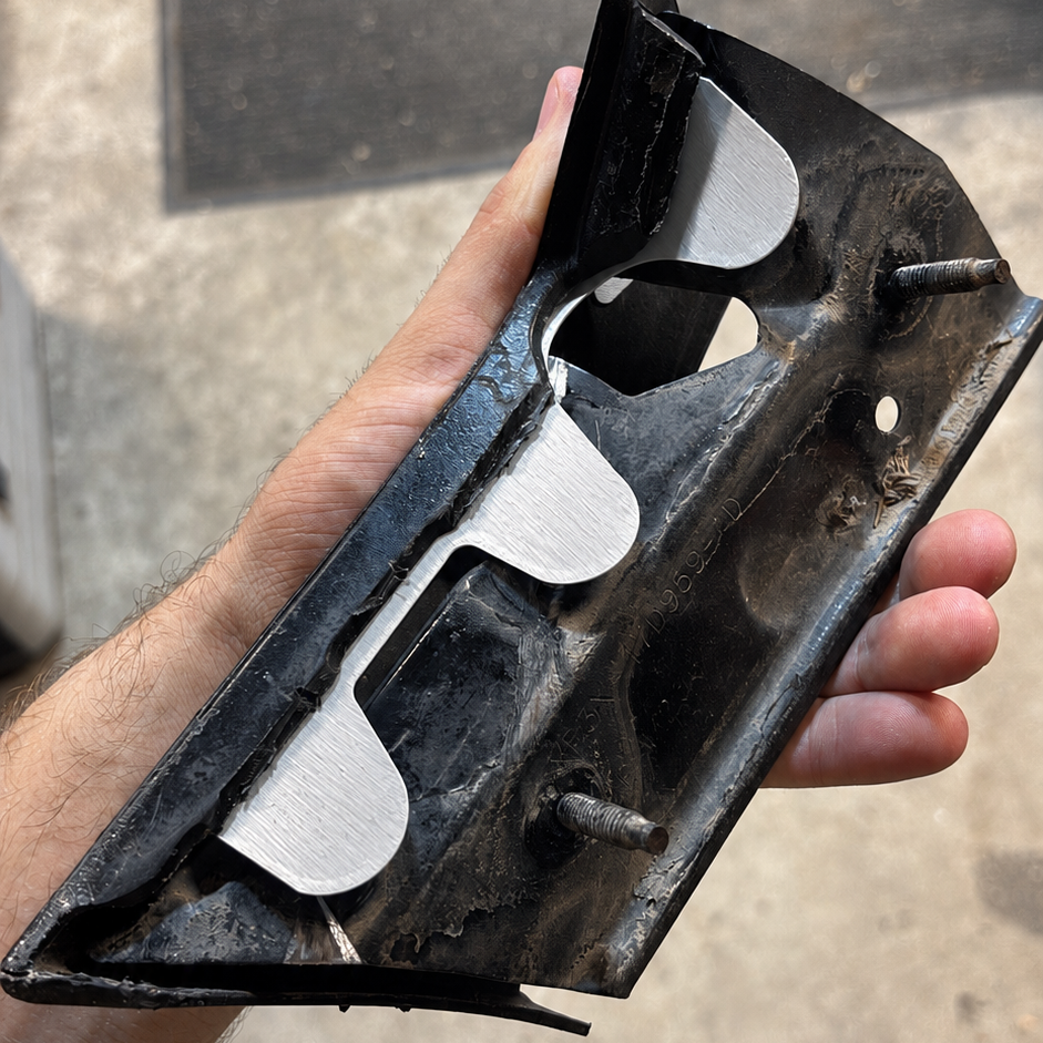A hand holding a black metal automotive part, likely a bumper bracket or trim piece, with silver-colored mounting tabs and screws, against an outdoor concrete background.