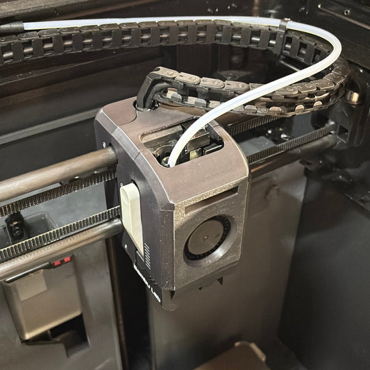 A close-up view of a 3D printer's print head assembly with a white filament cable, inside a black enclosure. A spool of blue filament is visible in the foreground.